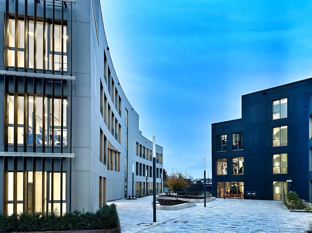 Gepflasterter Innenhof zwischen zwei Bürogebäuden in der Dämmerung, mit Leuchtenmasten und einem eingefassten Pflanzbeet mit Baum