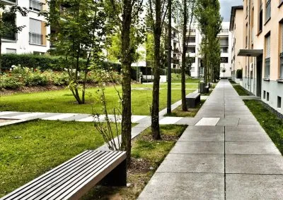 Breiter Plattenweg entlang eines Wohnhauses, daneben eine Rasenfläche mit Bäumen, Holzbänken und einem Abfallbehälter