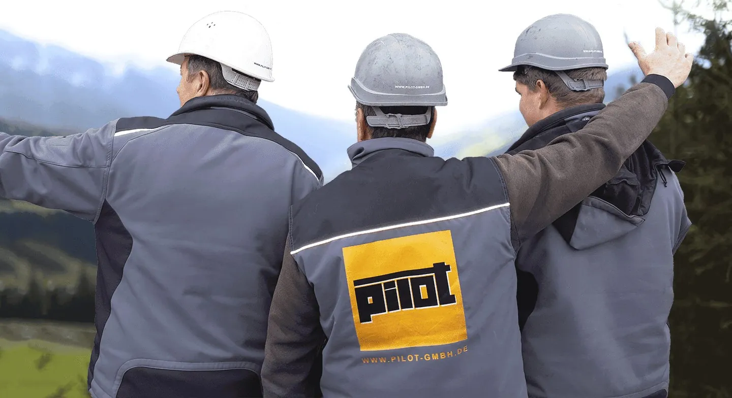 Drei Mitarbeiter der PILOT GmbH von hinten mit Schutzhelmen und Arbeitsjacken mit PILOT-Zeichen, zwei zeigen in die Landschaft