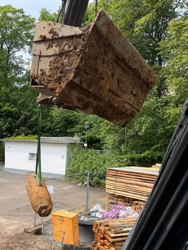 Blick aus der Baggerkabine: Eine verrostete Fliegerbombe hängt an einem grünen Hebegurt unter der erdverschmierten Baggerschaufel über der Baustelle.