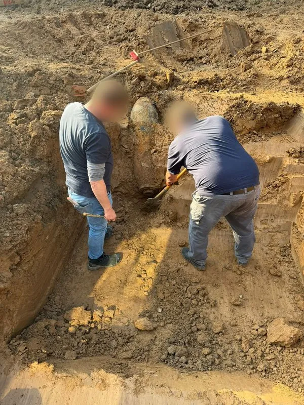 Zwei Männer in dunklen Poloshirts stehen in einer sandigen Baugrube und legen mit Spaten und Sonde eine verrostete Fliegerbombe frei, die schräg mit der Spitze nach oben im Erdreich steckt. Gesichter unkenntlich gemacht.