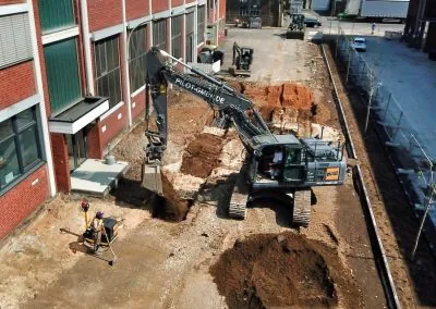 Kettenbagger der PILOT GmbH hebt zwischen zwei Gebäuden einen Leitungsgraben aus, daneben misst ein Mitarbeiter mit der Messlatte ein