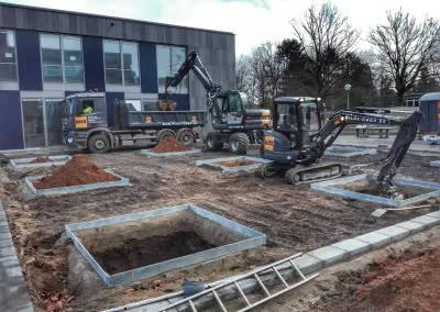 Ausgehobene quadratische Baumgruben mit Stahleinfassung auf der Schulhofbaustelle, dahinter zwei Bagger und ein Kipplaster