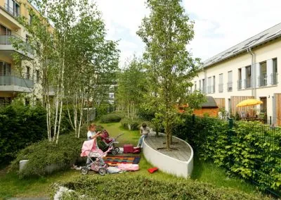 Begrünter Innenhof zwischen Wohnhäusern mit Birken und Rasen, auf einem geschwungenen Betonelement mit Holzbelag spielen zwei Kinder