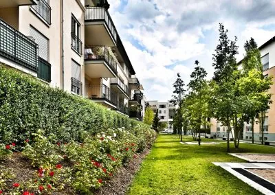 Rosenbeet und geschnittene Hecke entlang der Balkonfassade eines Wohnhauses, daneben eine Rasenfläche mit Bäumen und Plattenweg
