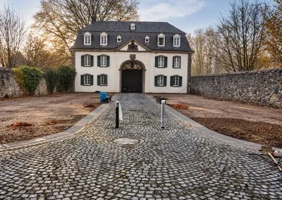 Gepflasterte Zufahrt im Bogenverband führt auf ein historisches Haus mit Rundbogentor zu, seitlich offene Erdflächen