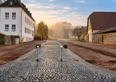 Gepflasterter Weg mit zwei Pollern zwischen einem hellen Wohngebäude und einem Wirtschaftsgebäude im Nebel
