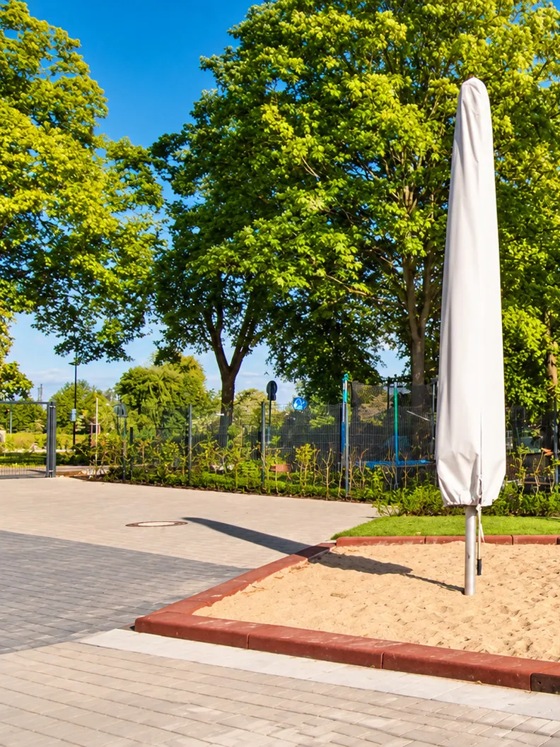 Sandkasten mit roter Einfassung, Sonnenschirm, Rasen und grauen Pflasterflächen vor einem Backsteingebäude unter alten Bäumen