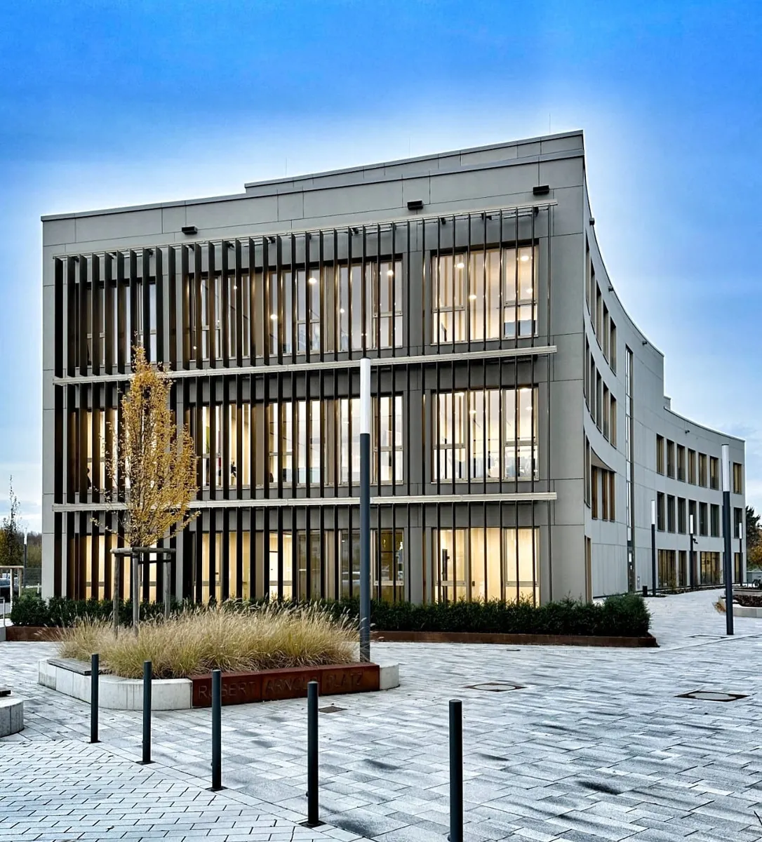 Beleuchtete Lamellenfassade eines Bürogebäudes am Bahnhof Köln-Wahn am Abend, im Vordergrund Ziergräser