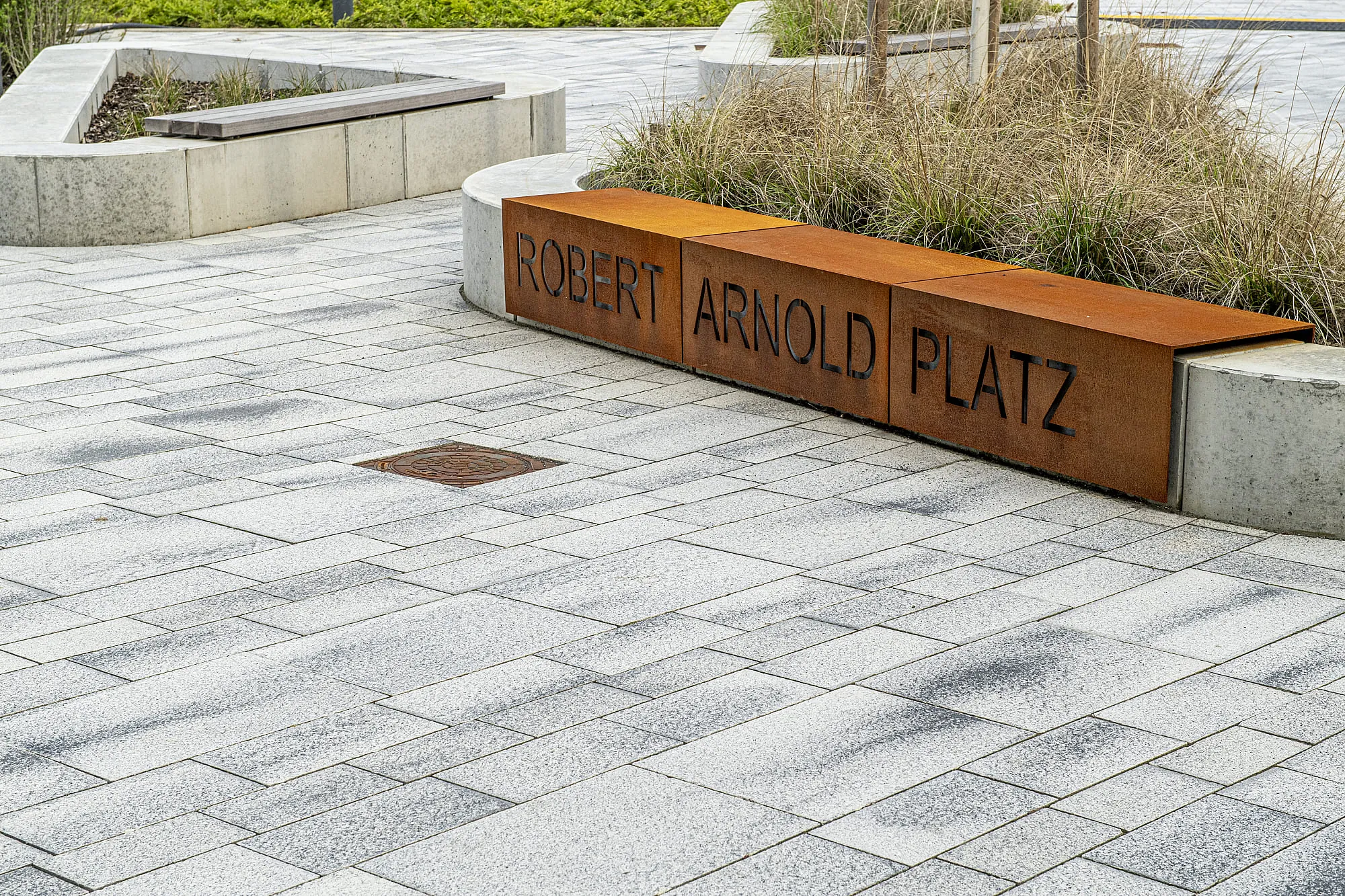 Gepflasterter Platz mit Betoneinfassungen und Gräserbeeten, eine Bank aus Cortenstahl trägt den Schriftzug Robert Arnold Platz