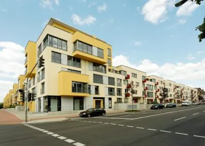 Asphaltierte Straße mit Gehweg und Fußgängerüberweg entlang einer neuen Wohnbebauung, am Fahrbahnrand parkende Autos und junge Bäume