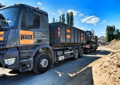 Lastwagen der PILOT GmbH mit Abrollcontainer auf einer Baustelle, dahinter ein Tieflader mit Bagger neben aufgeschüttetem Sand