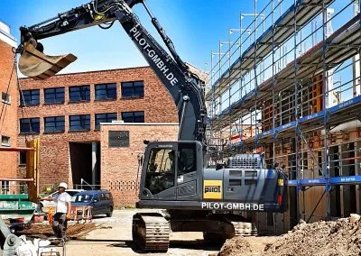 Kettenbagger der PILOT GmbH vor einem Backsteingebäude mit Baugerüst, links arbeitet ein Mitarbeiter an der offenen Baugrube